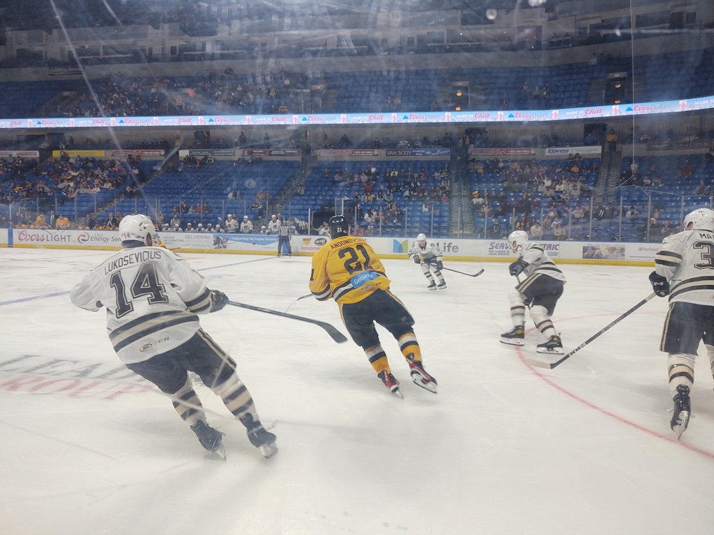 Image of game action on the ice at a hockey game. One player in a yellow jersey is surrounded by players in white jerseys from the opposing team.