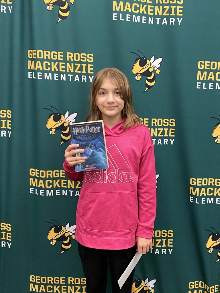 Charlotte Giglio, Grade 5, is standing in front of a green George Ross Mackenzie Elementary School backdrop. Charlotte is wearing a pink sweatshirt and black pants and is holding a Harry Potter  book.