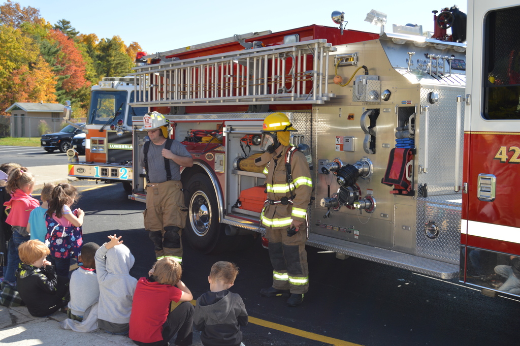 Two firefighters dressed in their uniforms stand in front of a fire truck while elementary school students sit on a sidewalk in front of them.
