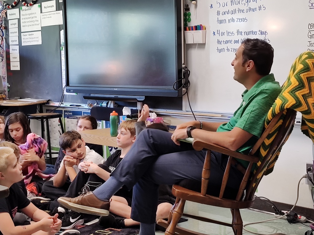 Image of building principal, Mr. Carpentieri, in a classroom, sitting in a rocking chair reading to students.