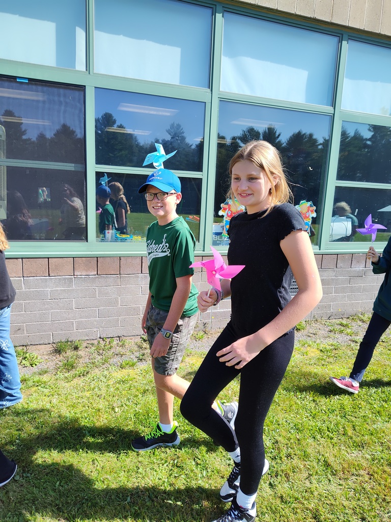 a student wearing a blue hat with a blue pinwheel stuck in it and another student with a pink pinwheel in her hand parade in front of a school building