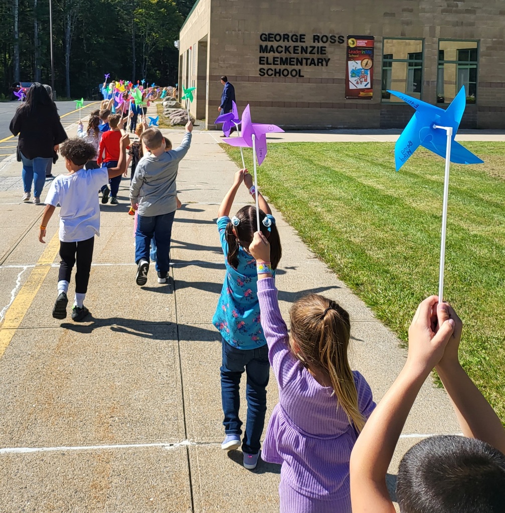 Image of the back of children as they walk past the camera holding their colorful pinwheels in the air parade in front of a school building.